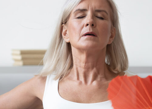 Photo of an older woman dealing with hot flashes, fanning herself inside
